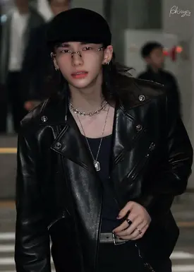 a young man with long, dark hair is captured in a closeup shot. he is wearing a black newsboy cap, thinframed glasses, a black leather jacket, multiple silver necklaces, and rings on his fingers.  his makeup is subtle, featuring dark lipstick. he appears to be posing for a photo, projecting a cool and edgy vibe. the background is blurred but suggests an urban, possibly nighttime setting. the overall aesthetic is moody and stylish. the image has a professional quality and evokes a sense of fashion or possibly a kpop idol aesthetic.