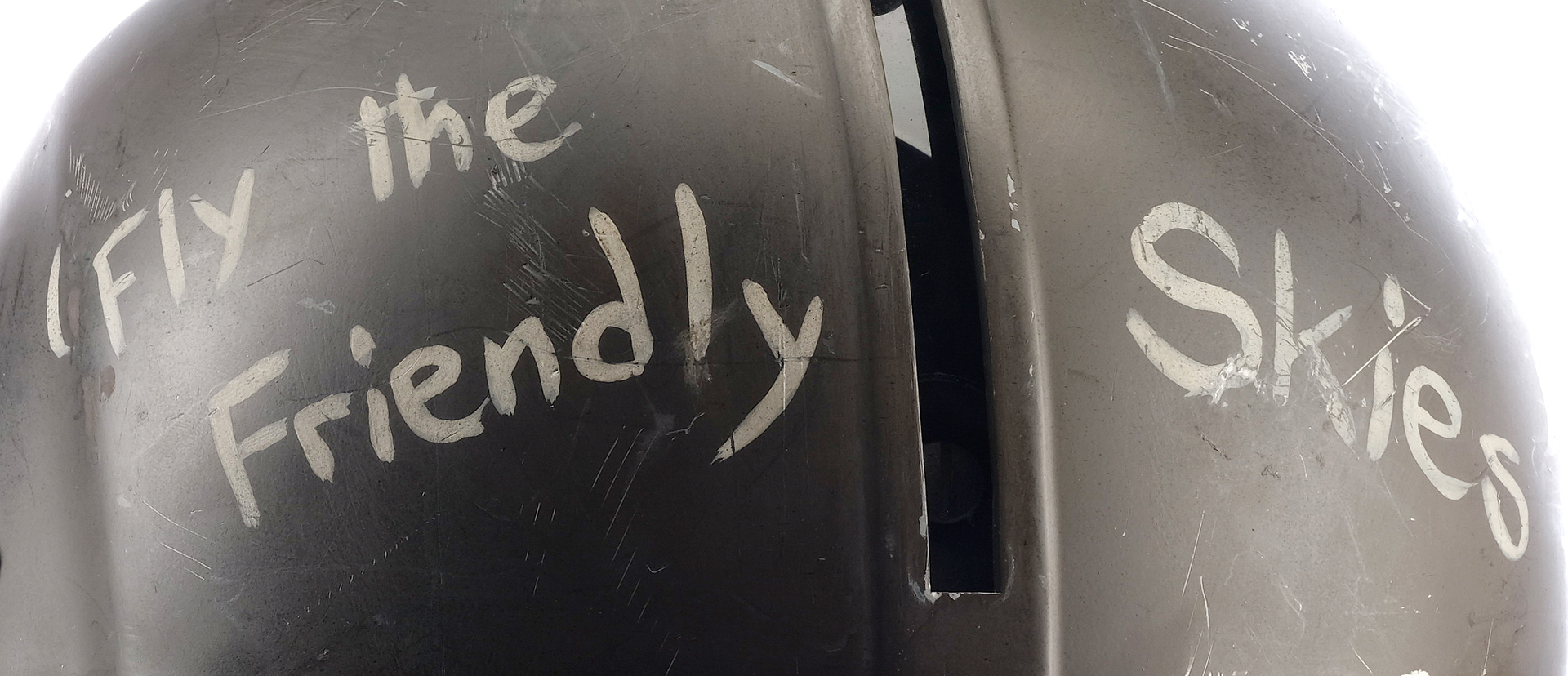 a closeup of a vintage military helmet with a worn, dented, and scratched exterior.  written on the side of the helmet in white chalk are the words, "fly the friendly skies," and other illegible writing.