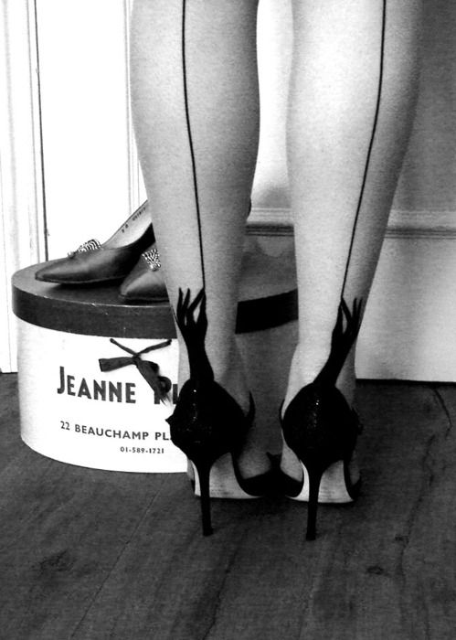 a black and white photo of a woman's legs and feet, wearing high heels with a back seam, standing next to a "jeanne r" shoebox.