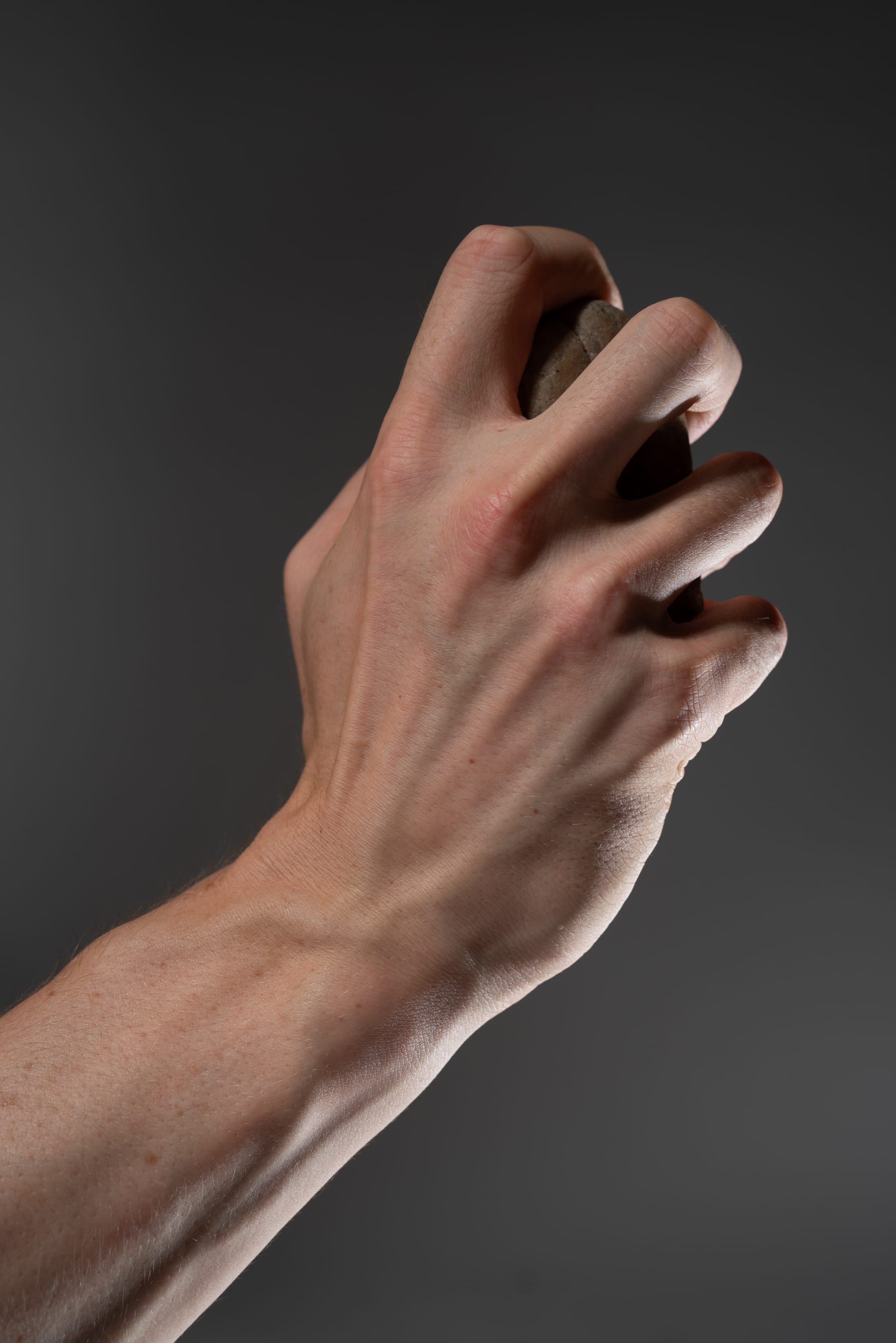 a closeup shot of a hand tightly gripping a rock against a dark gray background. the image emphasizes the strength and tension in the hand, highlighting details of tendons and veins.