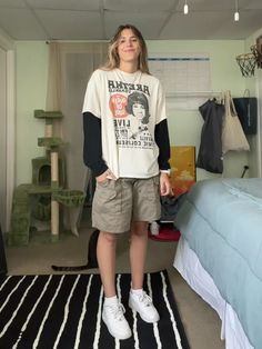 a young woman is standing on a black and white striped rug, in a room with a couch, a black and white striped wall, a cat in the background, and smiling at the camera. she is wearing a white tshirt with black long sleeves, with a picture of a woman on the front, and black cargo shorts. she is also wearing white sneakers. the woman is smiling and has long hair. the room is well lit, and the woman is looking confident and stylish.