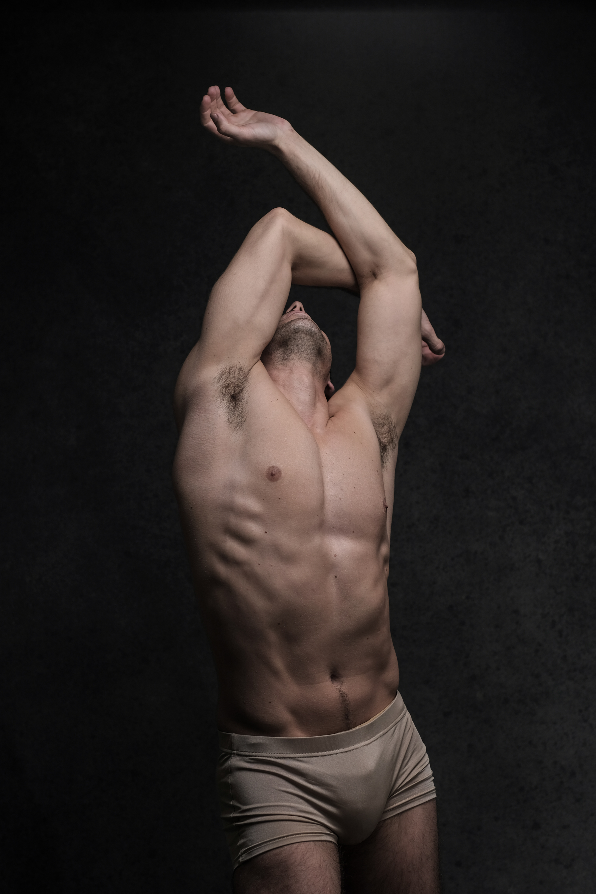 a muscular man stands in a dramatic pose against a dark gray background. he is wearing only underwear, and his physique is prominently displayed. the soft, lowkey lighting emphasizes his muscles and creates a sense of strength and athleticism. the perspective is slightly low, adding to the imposing nature of the image. the overall aesthetic is artistic and evocative, bordering on nude art.