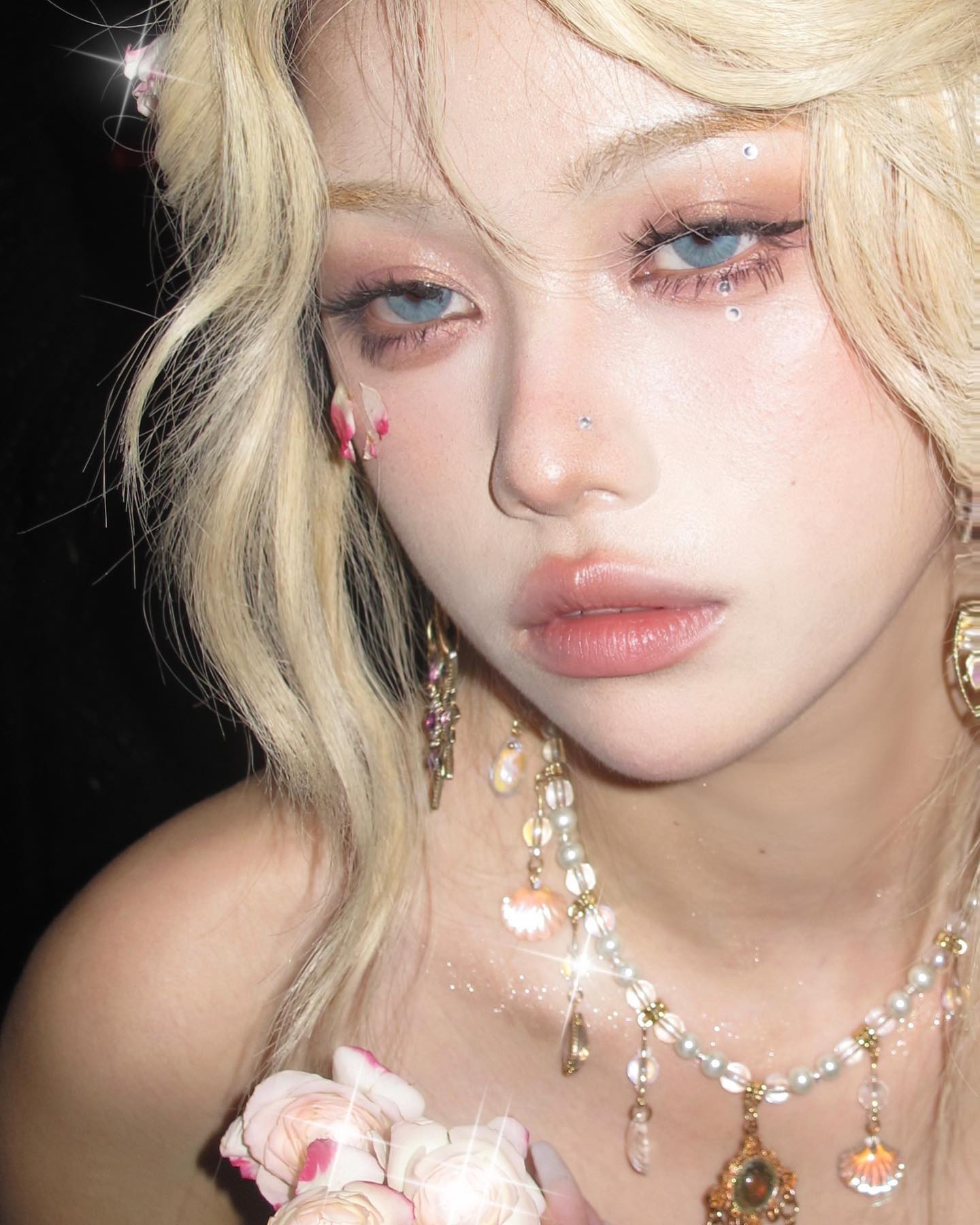 he image shows a closeup portrait of a young woman with blonde hair and pale skin. she has striking blue eyes enhanced with makeup and is wearing delicate, floralinspired makeup details. she is adorned with a pearl necklace featuring vintagestyle pendants and intricate earrings. the lighting is soft and artificial, creating a dreamy and ethereal atmosphere. the overall aesthetic of the image is romantic and feminine.