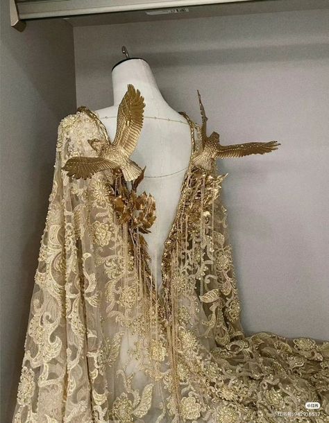 a goldcolored gown with intricate embroidery and lace is draped over a mannequin. the gown features two large, goldcolored birds perched on the shoulders.