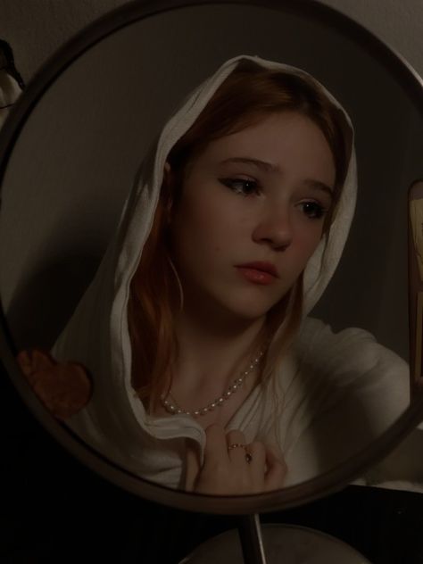 a woman with a white hood draped over her head is seen in a closeup reflection in a round mirror. she has a somber expression and is wearing a pearl necklace. the image has a vintage, cinematic feel with low, warm lighting.