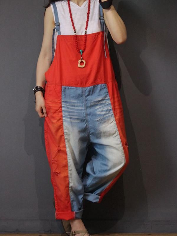 a woman is standing and leaning against a gray wall, wearing red and blue denim overalls over a white tshirt, a red necklace with a wooden pendant, a bracelet, and brown sandals. the overalls have a red top and sides, with blue denim in the center of the pants.