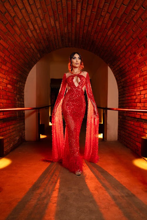 a woman in a striking red sequined gown stands in a bricklined tunnel. the dress features long, flowing sleeves with cutouts, adding to its glamorous and elegant design. the woman is adorned with jewelry, including earrings, which complements the attire. the tunnel's brick architecture and strategic lighting cast shadows on the walkway, enhancing the scene's overall ambiance. the dress seems to be a perfect choice for evening wear.