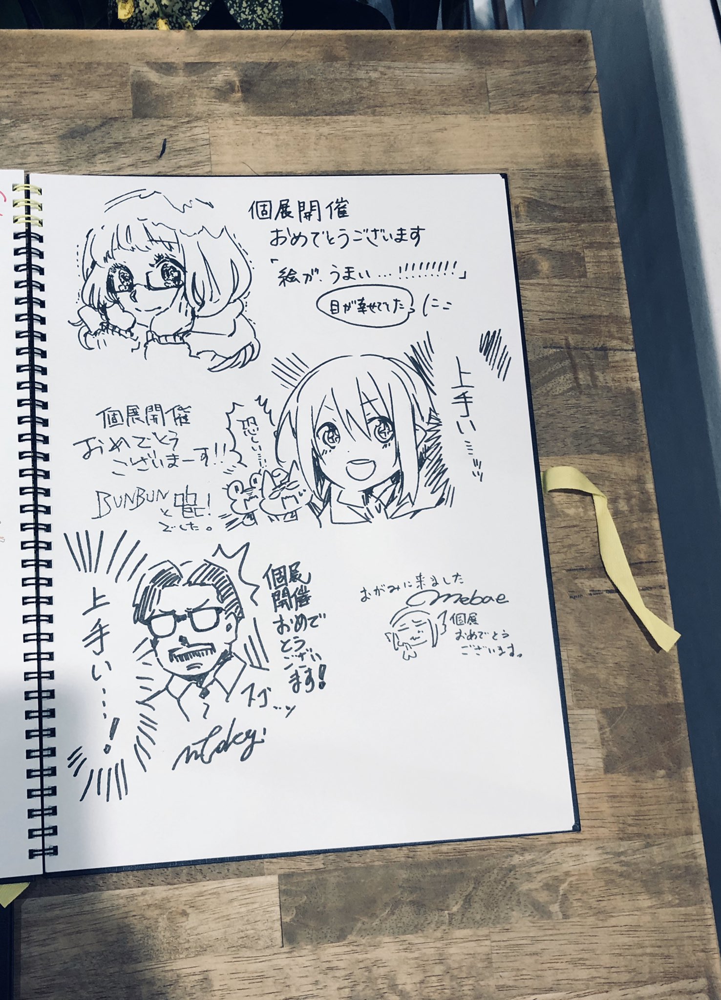 his is a photo of a sketchbook open to a page with four different anime style character drawings on it. each drawing has accompanying japanese text. based on the text, this page appears to be a collection of well wishes to the artist of the sketchbook on the occasion of their art exhibition. the book itself is sitting on top of a table that has a woodlike finish.