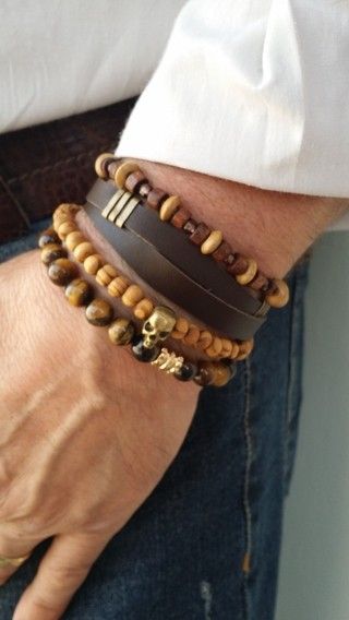 a man's arm wearing a leather bracelet with wooden and beaded bracelets on top.  the man is wearing blue jeans and a white shirt.  the bracelets are stacked on the man's wrist, making a stylish and layered look.