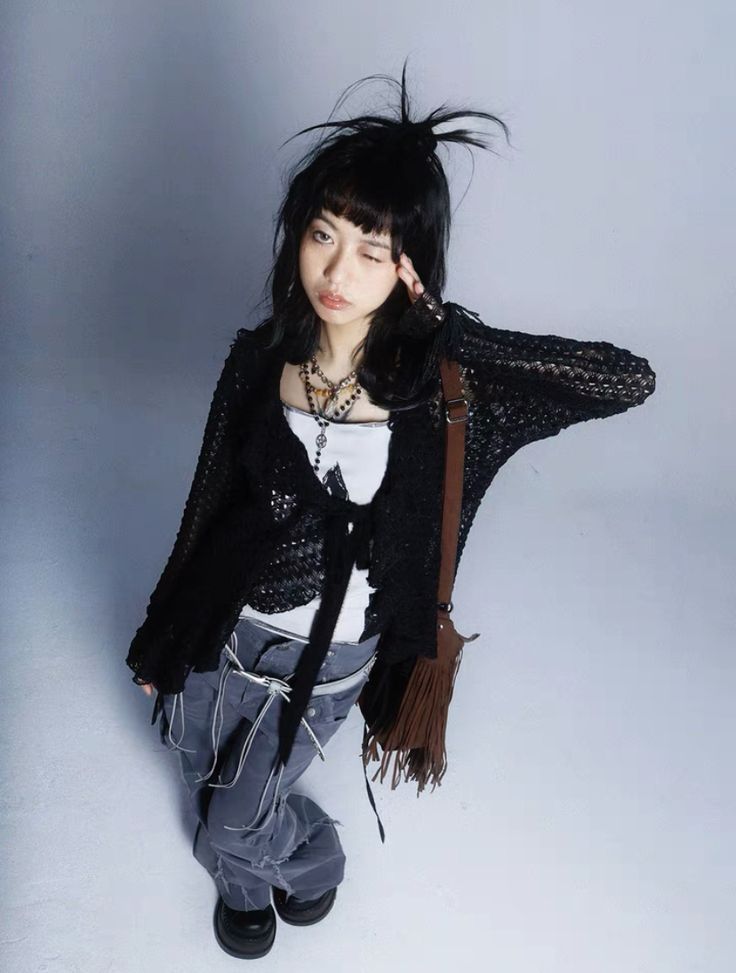 a highangle, fullbody shot of a young woman with dark hair styled in a slightly messy, highup bun posing against a plain white background. she is wearing a black openknit cardigan over a white top and grey distressed, baggy jeans. she also has on chunky black platform boots and carries a brown crossbody bag with fringe details.  her makeup includes eyeliner, and she has several necklaces. her overall style is alternative, incorporating elements of grunge, goth, and streetwear. the image appears to be a fashion photograph or a promotional shot.