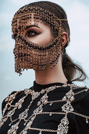 a woman is posing for a closeup portrait. she is wearing a glamorous, ornate gold and beaded mask that covers most of her face. her long dark hair is styled and she has a very stylish, elegant look about her.