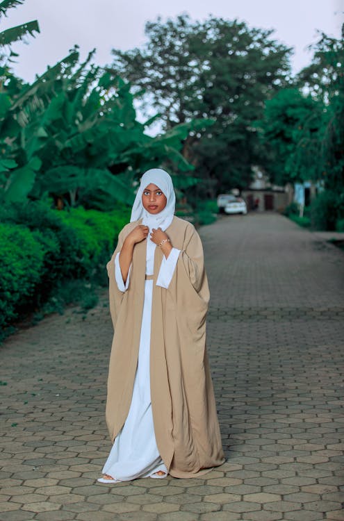 a fullbody portrait of a young woman wearing a beige abaya over a white maxi dress. she is also wearing a white hijab.  she is standing on a paved pathway bordered by lush green vegetation and trees, with a calm and serene expression on her face.  the overall mood is peaceful and elegant. the image is welllit and captures the details of her clothing and the surrounding environment.
