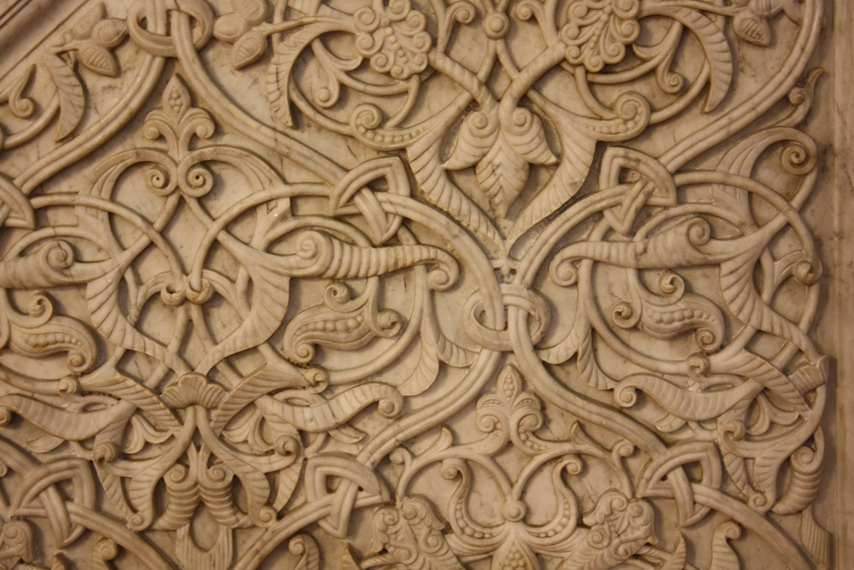 the image shows a detailed, closeup view of an intricately carved stone surface, possibly marble or a similar material. the carving features a complex ornamental pattern composed of floral and vinelike elements, scrolls, and leaf motifs. the texture is rich with depth created by the relief carving. the overall design is elaborate and visually captivating, showcasing the skill of the artisan.