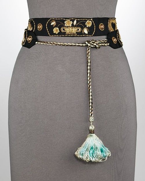 a black fabric belt with embroidered gold floral designs is displayed on a gray mannequin. the belt features a knotted gold cord that extends down with a white and teal tassel. the overall image has a clean and welllit studio style.