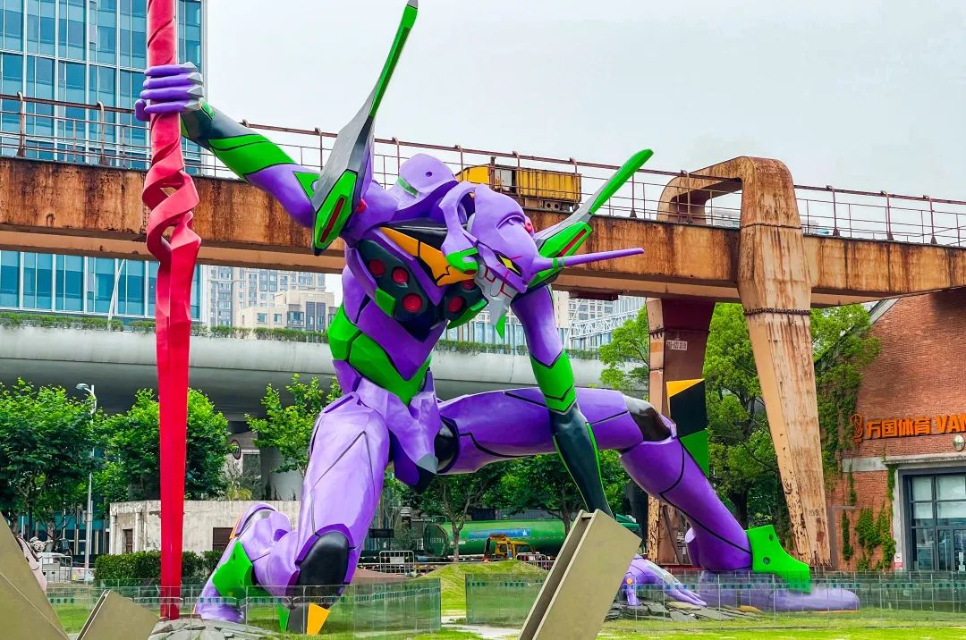 a large purple and green statue of the eva unit 01 from the anime neon genesis evangelion stands in a city park. the statue is posed in a dynamic stance, with its arms outstretched and its legs bent. it appears to be holding a large red spear in its right hand. the statue is set against a backdrop of tall buildings and a rusty metal structure. there are trees and other plants in the foreground. the sky is overcast and the air is still. the statue is a popular tourist attraction in the city.