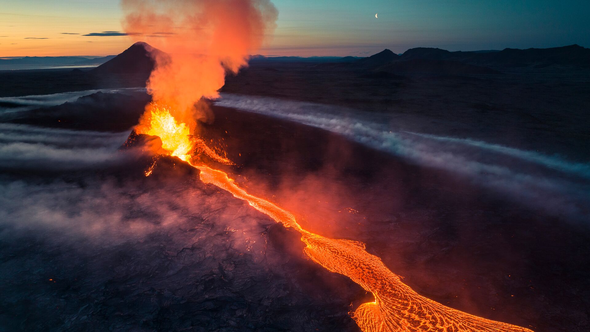 an aerial view of a volcanic eruption at night. the bright orange lava flows down the side of the mountain, creating a dramatic and fiery scene. the sky is dark and cloudy, with a crescent moon visible in the upper left corner. the image is a powerful reminder of the beauty and destructive power of nature.
