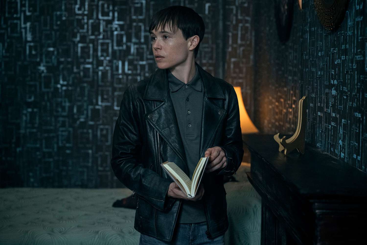 elliot page as vanya hargreeves in the umbrella academy, stands in a bedroom, holding a book and looking to the side. the room is dimly lit with a blueish tone, and the walls are decorated with a geometric pattern.  the overall mood is mysterious and contemplative.
