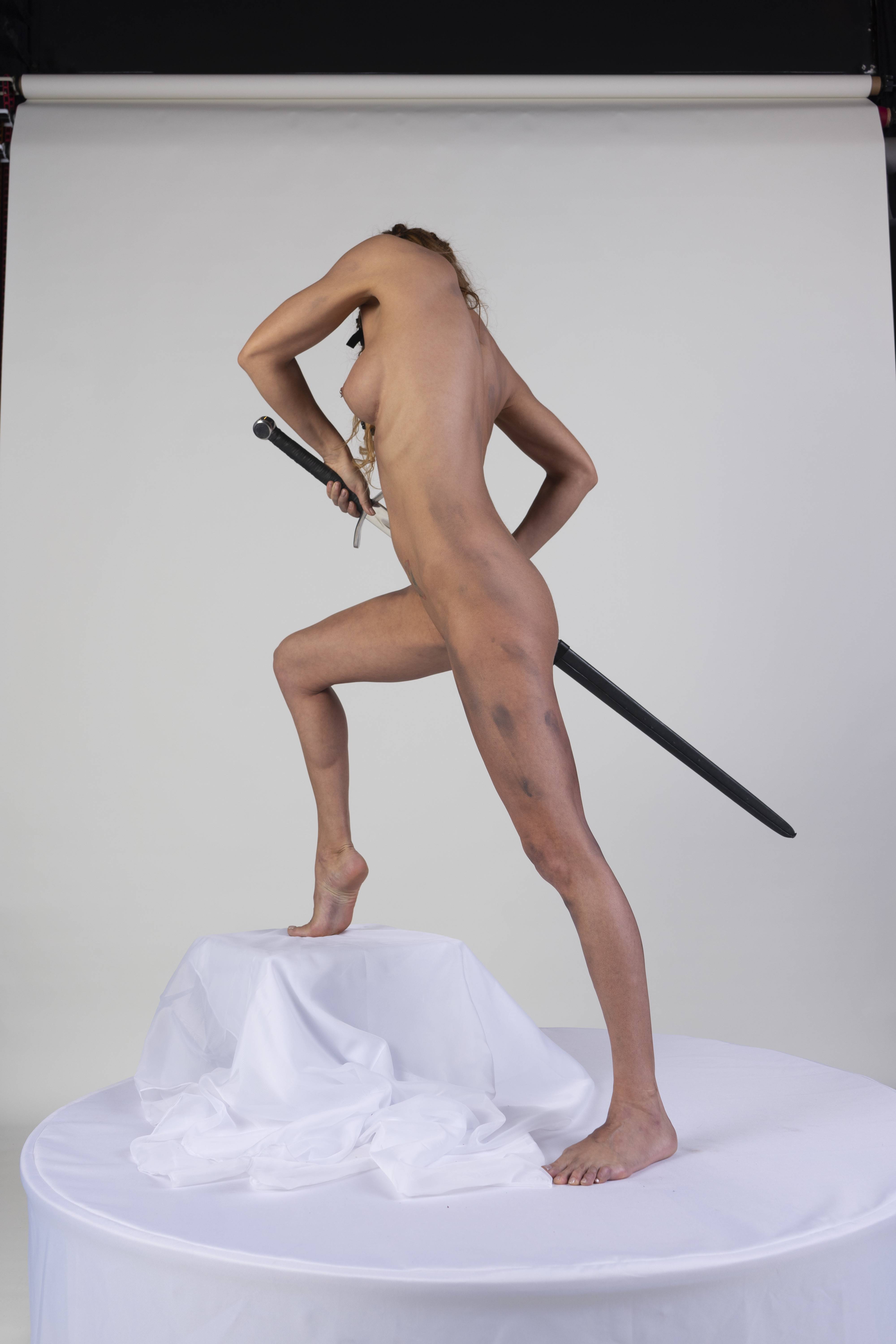 a nude woman stands on a white table, holding a sword. she has bruises on her body. the background is a white studio backdrop.
