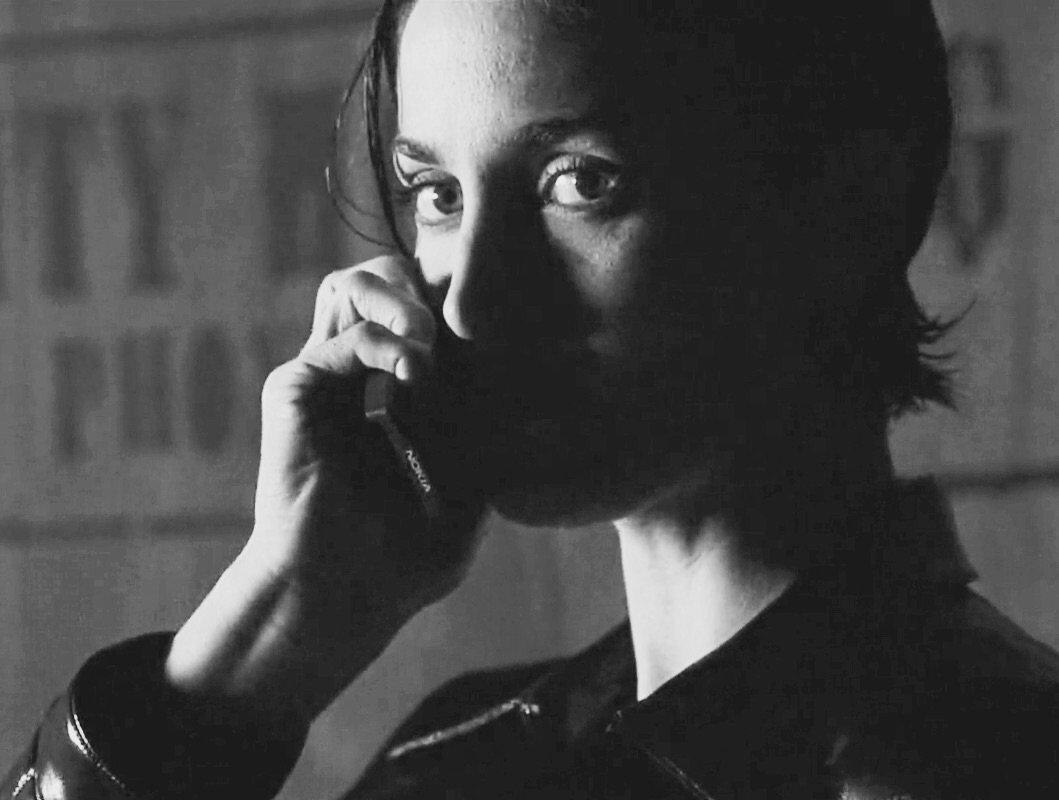 the image is a black and white closeup portrait of a woman. she has short, dark hair and is holding a telephone to her ear with her hand partially obscuring her face.  she's wearing a darkcolored, possibly leather, jacket. the background is blurred and out of focus, featuring parts of what looks like a wall with faded lettering. the overall mood is dark, moody, and slightly mysterious, with strong contrasts of light and shadow accentuating her features and expression.  the woman's gaze is somewhat intense and pensive.