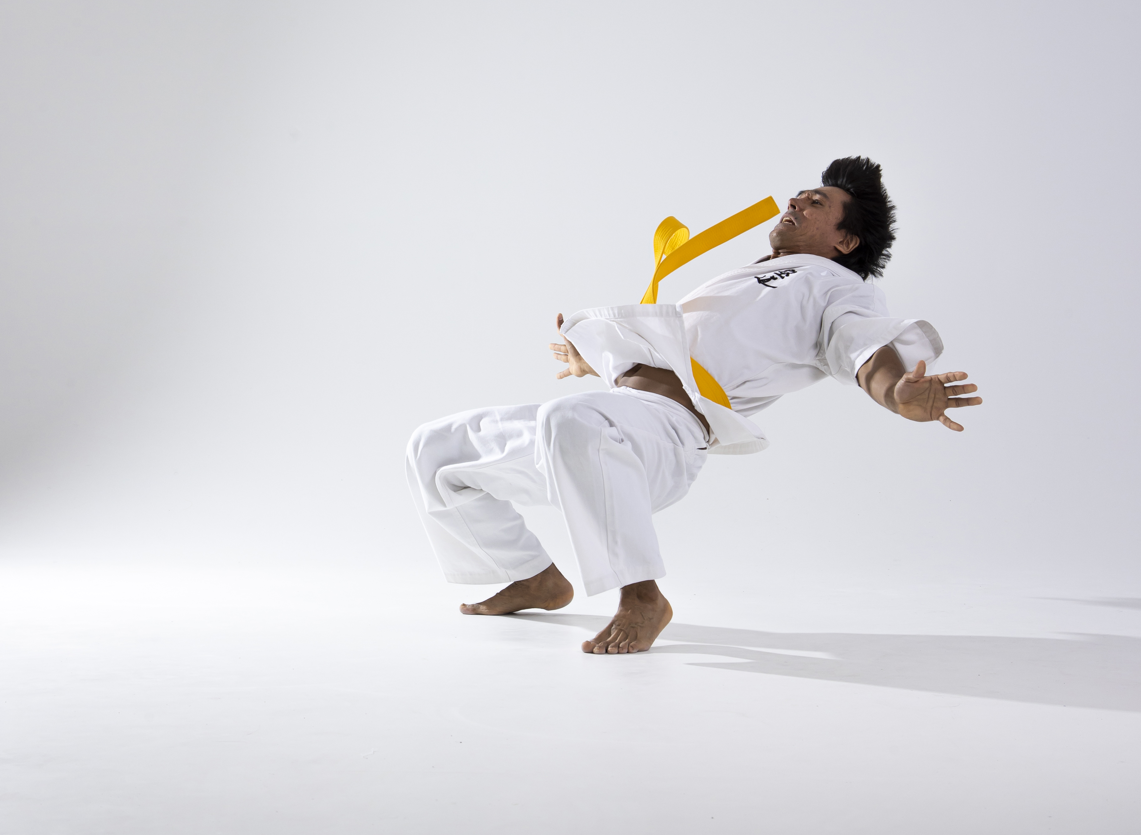 a man in a white karate uniform with a yellow belt is performing a dynamic jumping movement. he is barefoot, and the photo is a studio shot against a light background. the man has dark hair and is in midair, capturing the athleticism and motion of the sport.