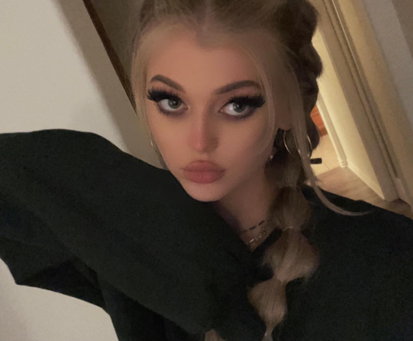 a closeup selfie of a woman with blonde hair styled in braids, pouting with large eyelashes and hoop earrings, wearing a black sweater indoors.