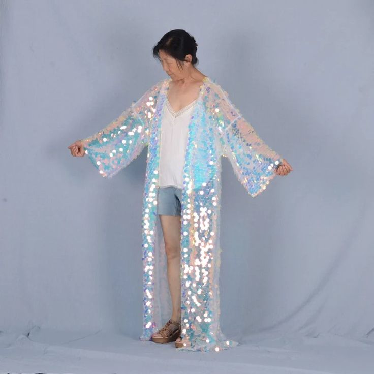 a woman in a white iridescent sequin kimono posing against a light blue background. the kimono has long sleeves and a flowing silhouette. she is wearing denim shorts and sandals underneath.  she is looking down and her hair is pulled back.  she is showing off the sequin details of the kimono.
