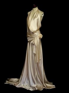 a white silk gown with a draped halter neckline. the gown is worn by a mannequin in a studio setting against a black background. the fabric is very smooth and elegant.