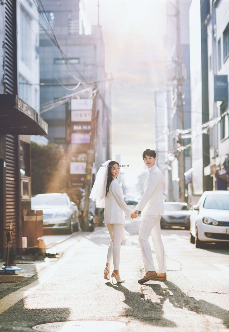 a young couple, dressed in white wedding attire, walk handinhand down a sundrenched city street. the bride wears a chic pantsuit and veil, while the groom sports a sharp suit. they are captured from behind, looking back at the camera with joyful expressions. the urban setting provides a modern and stylish backdrop for this romantic moment. the warm, natural light enhances the image's overall sense of happiness and celebration.