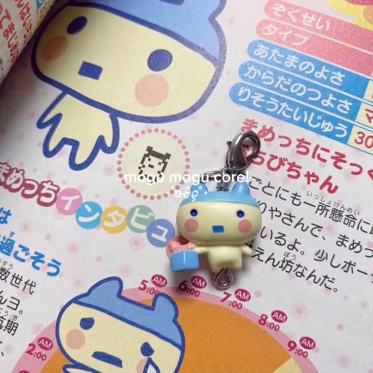 the image shows a book with cartoon images of tamagotchi characters and japanese text, with a tamagotchi charm hanging near the page.