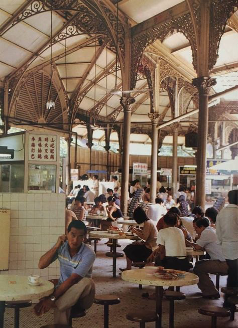 the image showcases the interior of a bustling food court, likely in singapore, during the 1970s or 1980s.  the architecture is striking, featuring a high ceiling supported by ornate castiron columns and arches.  the space is densely populated with people seated at numerous small tables, enjoying meals.  various food stalls are visible in the background.  the overall atmosphere is lively and characteristic of a traditional asian hawker center. a man in a light blue shirt is prominently seated in the foreground.  the style of the architecture suggests a colonialera building.  there are signs with chinese characters visible.