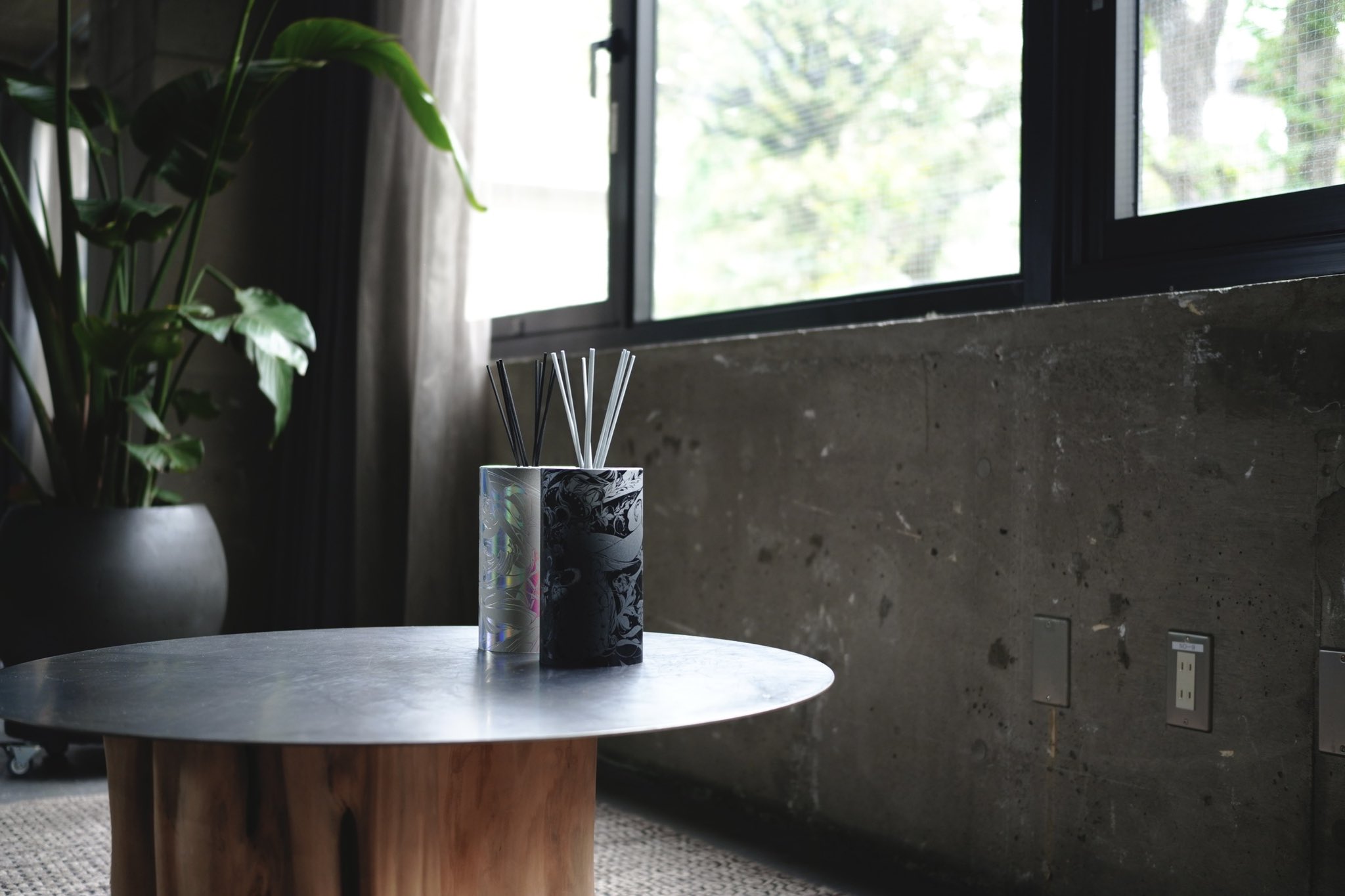 this is a photo of a living room. there is a large plant in a pot in the corner. in the center of the photo is a low, round table made of metal and wood. on top of the table sits a black and white diffuser. there is a large window in the background. the walls are concrete and the room is brightly lit with natural light.