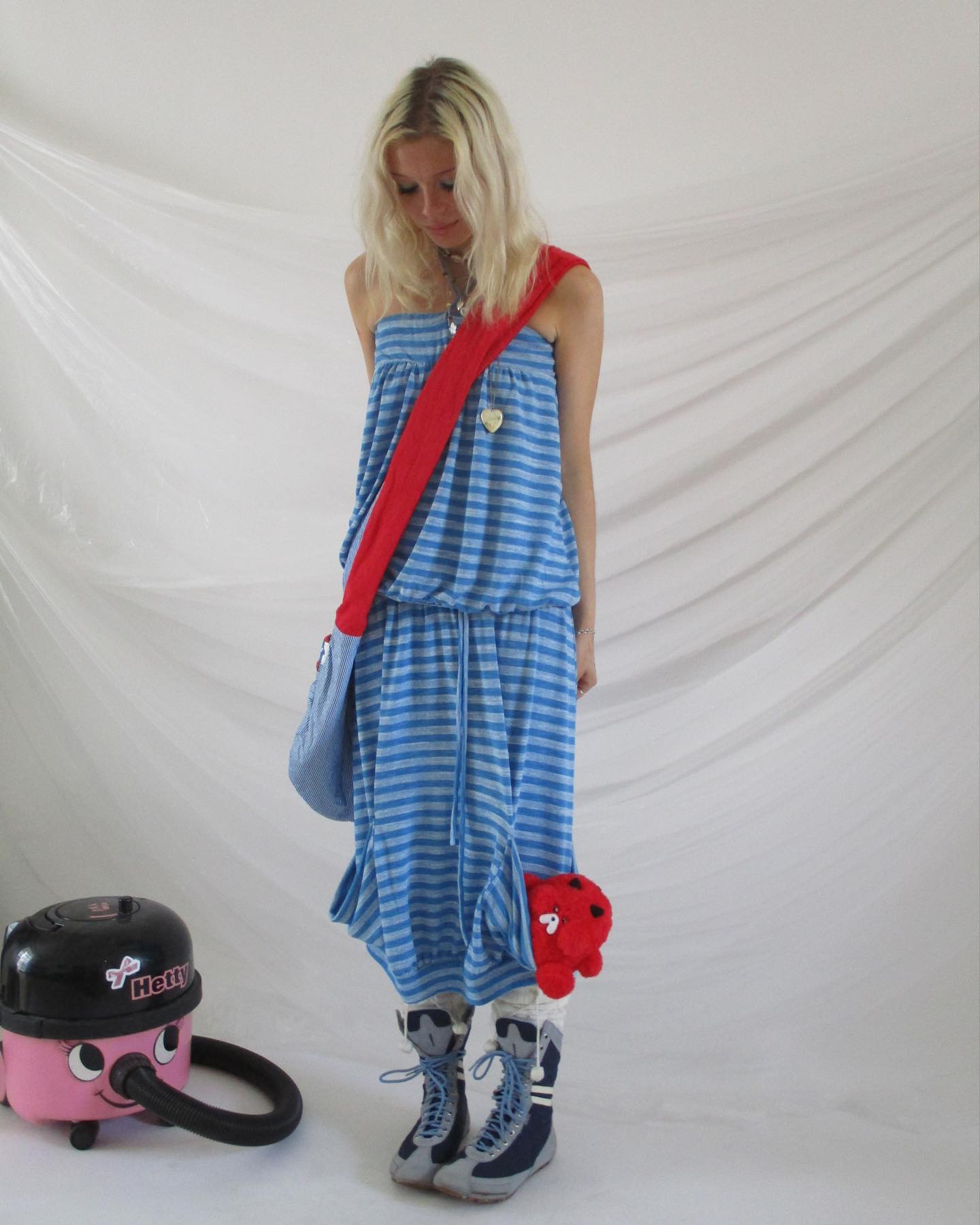 young blonde woman is standing in front of a white background. she's wearing a light blue and white striped maxi dress, a red fabric shoulder bag and high top boots. a small red toy is tucked into her dress pocket. a pink vacuum cleaner is on the floor near her feet.  the overall style is casual and slightly quirky.