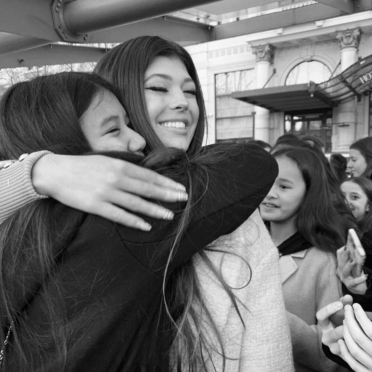 a black and white shot captures a woman joyfully embracing a young girl in what appears to be a public space. the woman is wearing a lightcolored coat, while the girl is bundled in a dark jacket. both are smiling, showing genuine emotion. other young girls are gathered in the background, some holding up phones, possibly as fans or onlookers. a building with the word "hotel" can be seen behind the group. the scene suggests a heartwarming encounter between the woman and her admirers.