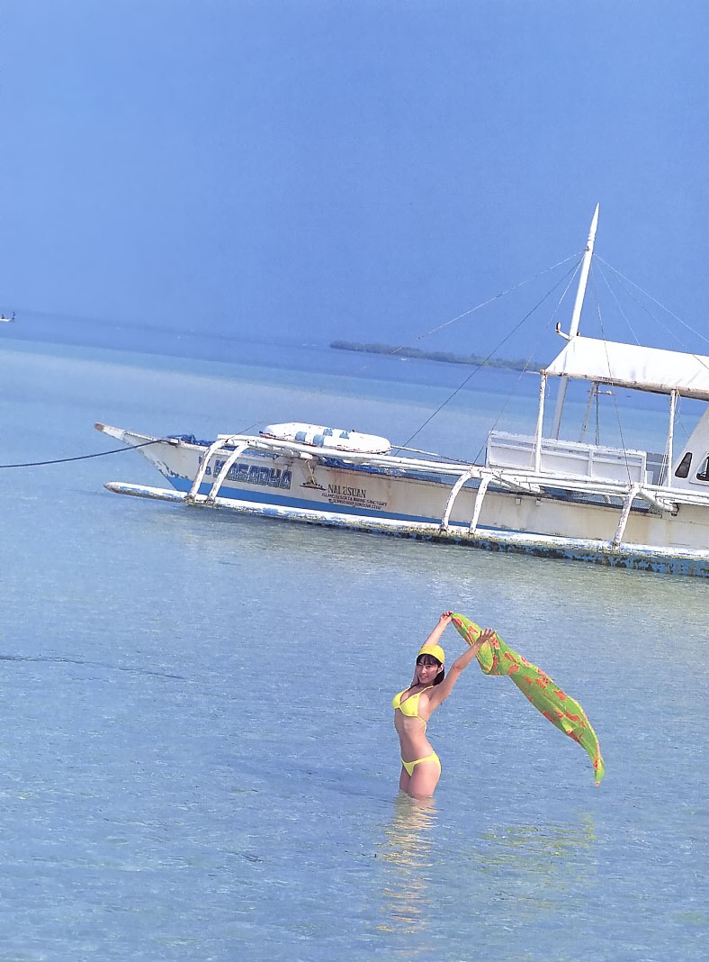 a woman in a yellow bikini stands in shallow, clear blue water, holding a green sarong that's billowing in the wind. a traditional philippine banca boat is anchored behind her. the background features a pristine beach and a clear sky, evoking a sense of tropical paradise.