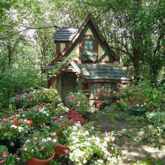 a small, charming cottage nestled in a lush garden setting within a forest. the cottage features a steeply pitched roof, dormer windows, and a welcoming front porch adorned with flower boxes. the surrounding garden is overflowing with colorful flowers in various shades of pink, red, and white. abundant greenery and mature trees create a sense of seclusion and tranquility.