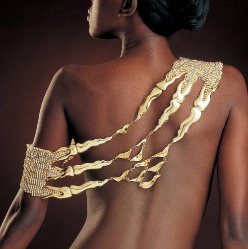 an image features a darkskinned woman from the back, showcasing a gold shoulder piece and arm bracelet. the shoulder piece is intricately designed, while the arm bracelet complements the overall fashion statement.