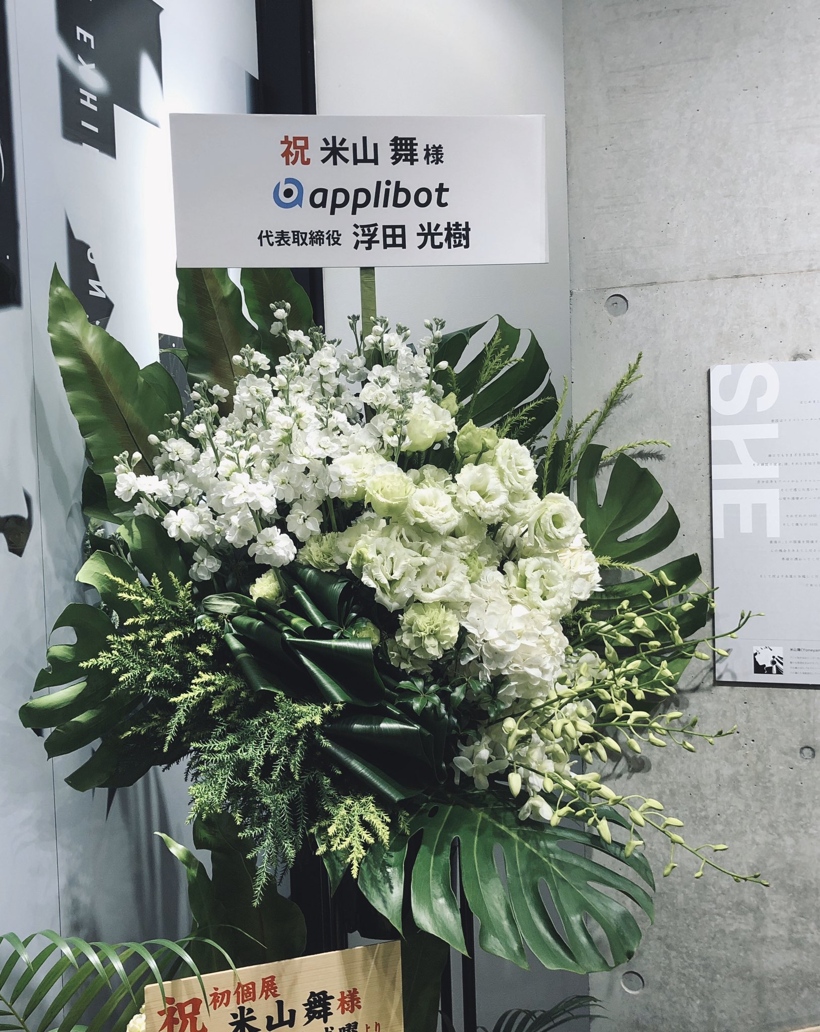 a large congratulatory floral arrangement, primarily white flowers with lush green foliage, stands before a gray wall adorned with a congratulatory message in japanese. the message celebrates a ms. mai koyama's first solo exhibition and features the name "applibot" with its ceo, mr. mitsuki ukita. the arrangement exudes elegance and celebration, signifying a momentous occasion.