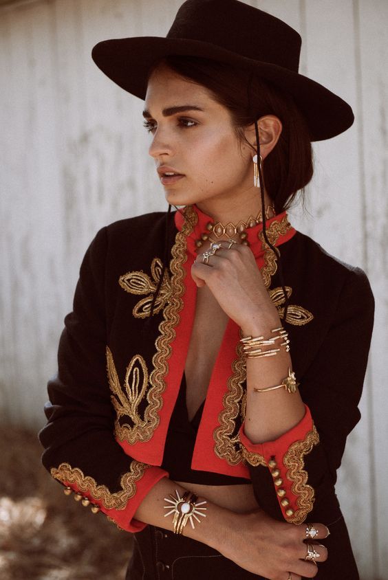 a woman wearing a black hat and an embroidered black and red jacket poses with her hands near her neck, showcasing multiple gold rings, bracelets, a choker, and earrings. she is looking off to the side.