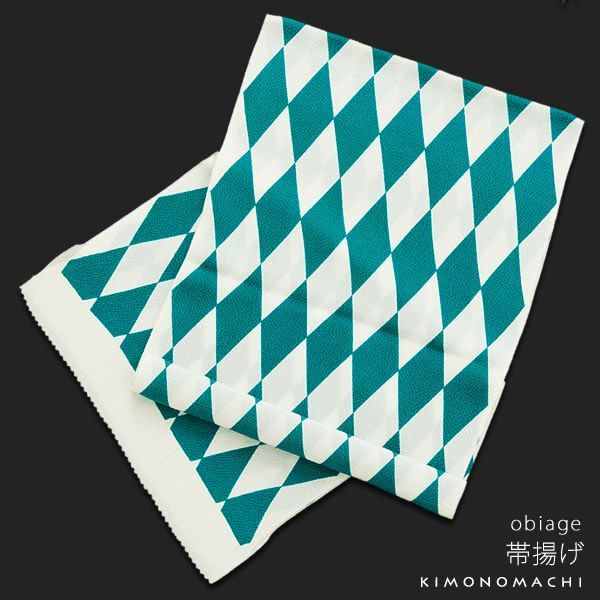 the image features a folded obiage, a traditional japanese kimono accessory, with a geometric diamond pattern in teal and white. the obiage is folded on a dark surface and has text printed in the lower right corner.