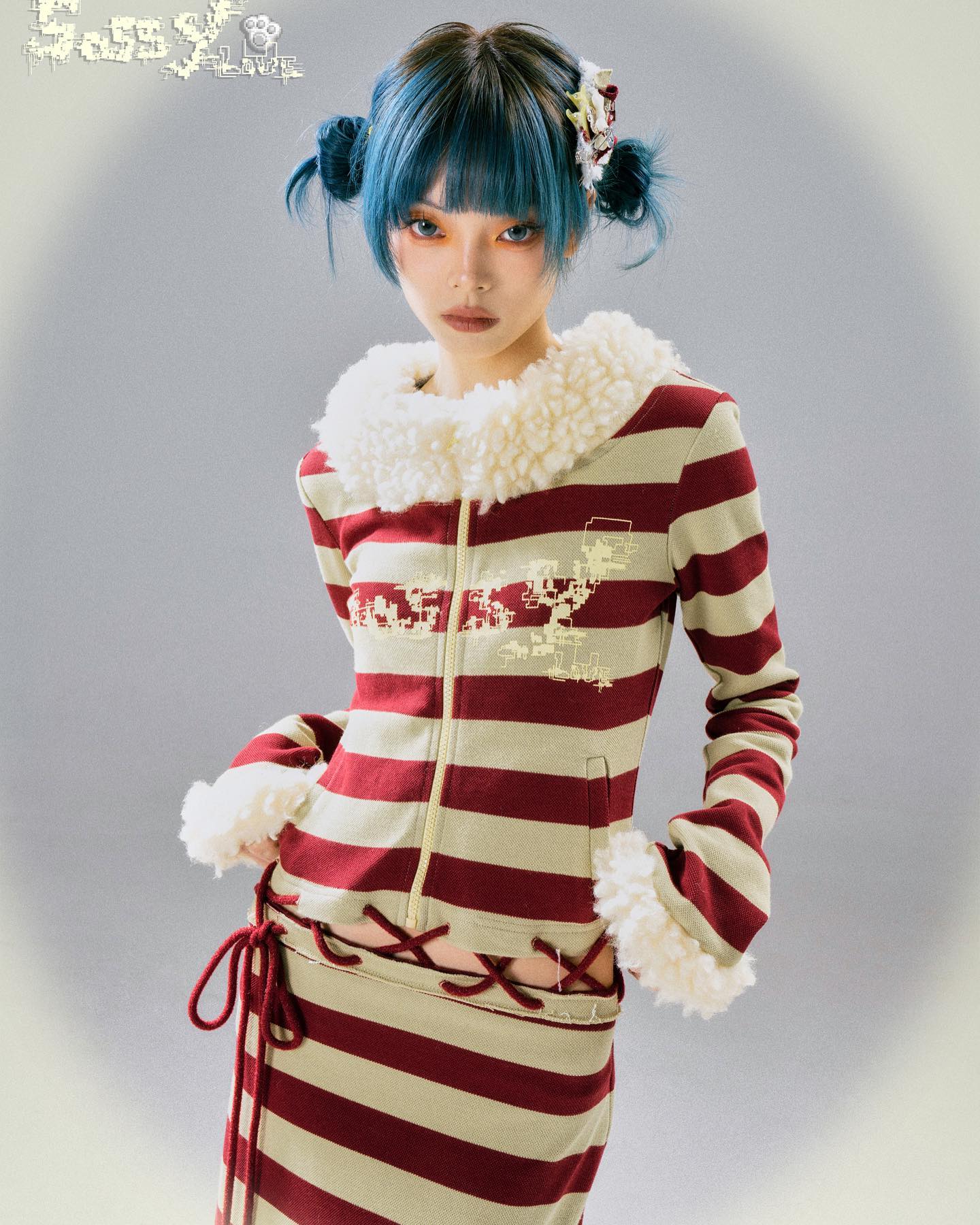 a young woman with vibrant blue hair styled in two buns, is showcased in a studio portrait. she's dressed in a striking ensemble: a red and beige horizontally striped zipup jacket with a fluffy white collar and cuffs, paired with a matching striped skirt. the skirt is cinched at the waist with crisscrossing laces. the overall style is reminiscent of anime or kawaii aesthetics. the brand "glossy lover" is subtly included in the image's watermark. the background is a neutral, muted tone that emphasizes the model and her outfit.