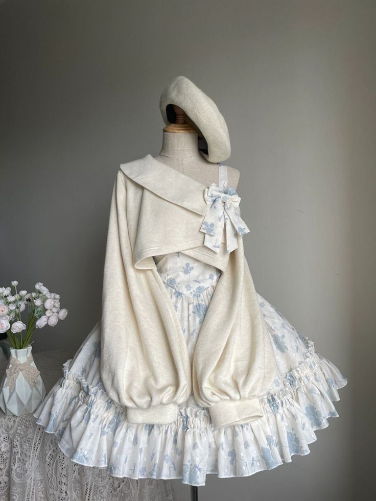 a delicate white and blue floral dress with an offtheshoulder cropped sweater is displayed on a mannequin. the sweater features dramatic oversized sleeves, a wide collar, and a blue bow on the shoulder. the skirt of the dress is full and flowing with multiple layers of fabric. the mannequin also wears a simple white beret tilted to the side.