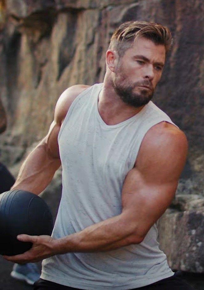 a muscular caucasian man with a beard is wearing a white tank top while holding a black exercise ball. he is looking away from the camera and smiling as he stands outside against a backdrop of rocks.