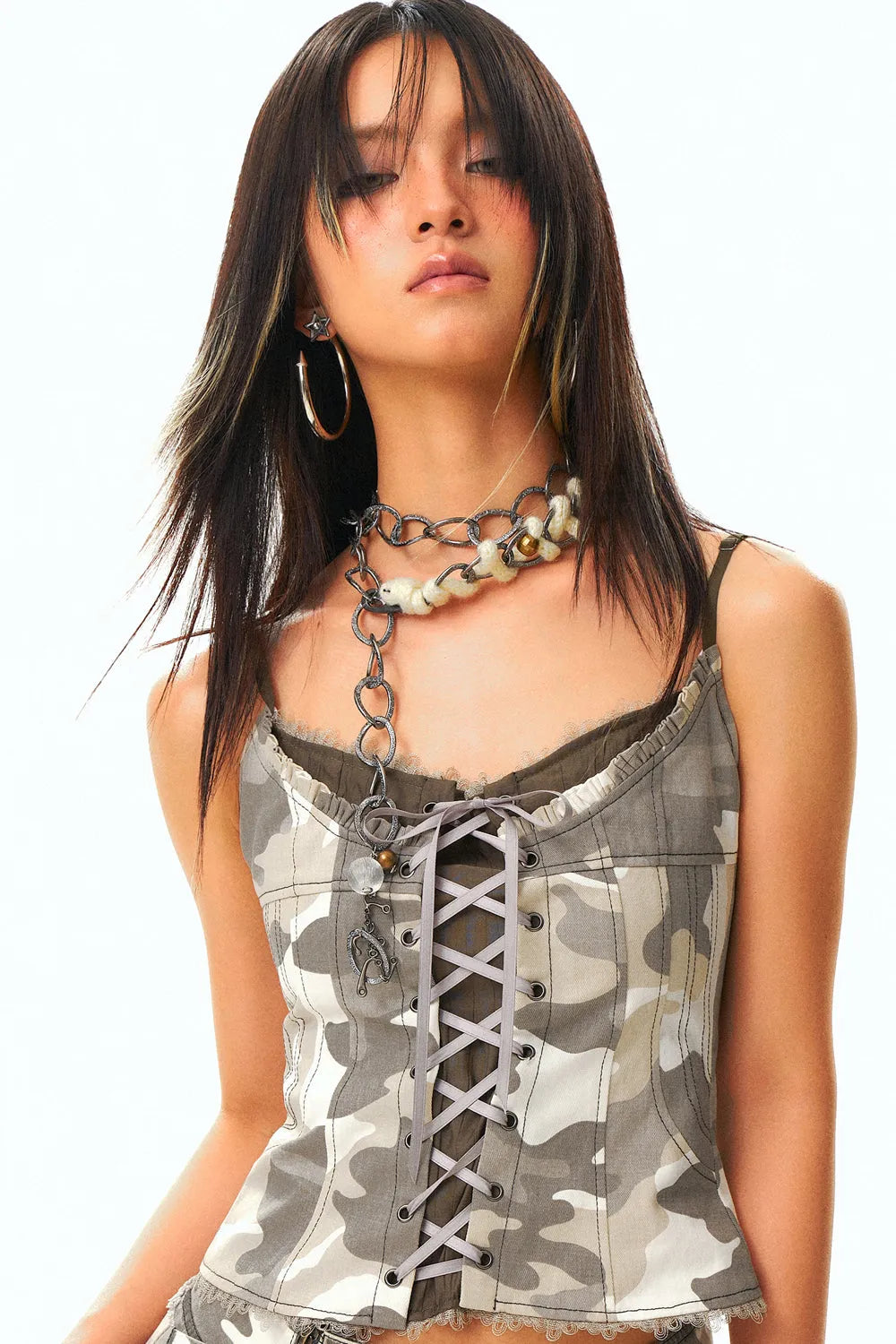 closeup portrait of a young asian woman with long dark hair styled with bangs and blonde highlights. she's wearing a light beige camouflage print corsetstyle tank top with a laceup front, layered over a dark brown camisole. she accessorizes with a layered necklace featuring a chain and beaded elements, and large hoop earrings. the overall style is trendy and youthful. the image is taken in a studio with a clean white background.