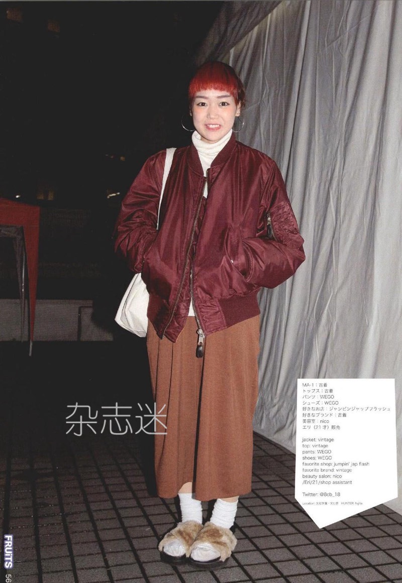 a young woman with bright red hair styled in a bun is captured in a lowlight, nighttime setting.  she embodies a trendy, casual style with a vintage feel. her outfit consists of a maroon bomber jacket, a beige turtleneck sweater, a brown midi skirt, white socks, and furry brown sandals.  she carries a white tote bag slung over her shoulder.  the background features a glimpse of an outdoor space with a white tentlike structure. the photo's aesthetic reflects late 90s/early 2000s japanese street fashion.  text in japanese, along with the word "fruits," appears at the bottom left of the image.