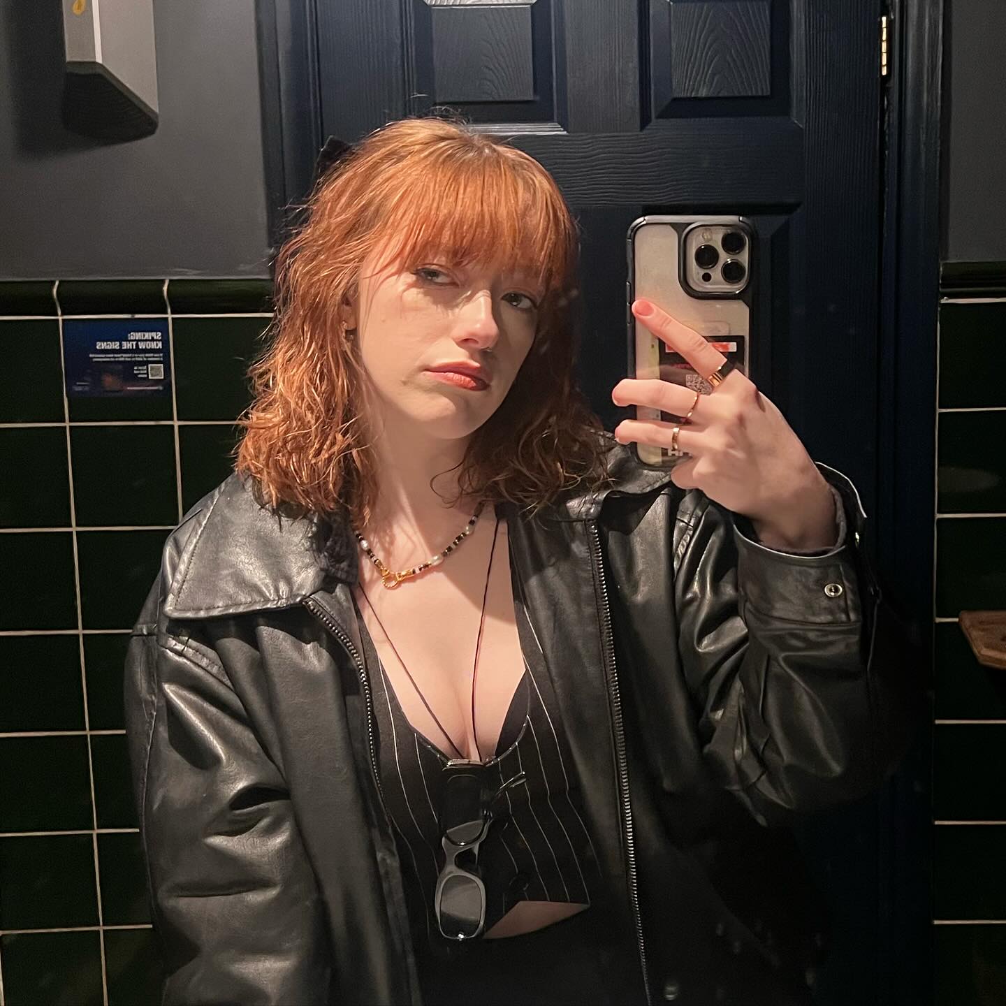 a young woman with vibrant red hair and bangs takes a selfie in a dark bathroom with green tiled walls. she's wearing a black leather jacket over a black and white pinstripe cropped top.  she's accessorized with a necklace and rings, and holds her iphone in one hand, making a peace sign with the other. the overall vibe is stylish and casual.