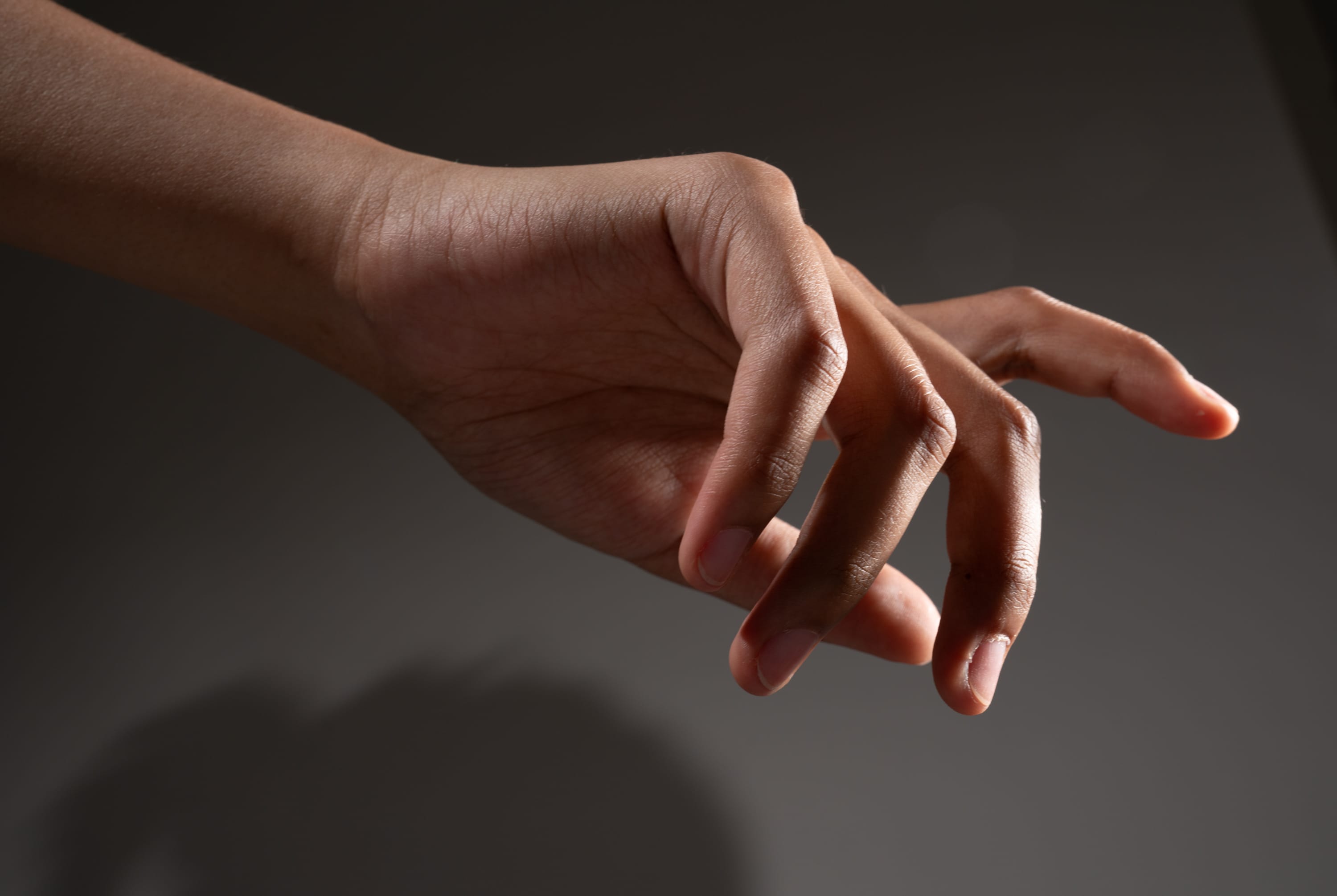 a hand is reaching out from the left side of the image with fingers slightly curled. the background is dark gray and the lighting is soft and dramatic. the hand has a warm skin tone.