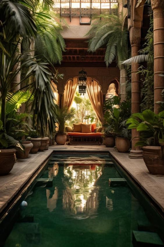 an indoor courtyard with a long rectangular pool in the center. lush green plants in pots line the sides of the pool, creating a tropical ambiance. the far end of the courtyard features a cushioned bench positioned under an arched doorway covered with flowing drapes, providing a glimpse of a sunlit garden beyond. the pool reflects the surrounding architecture and greenery, enhancing the sense of tranquility and serenity. the space is bathed in soft, warm natural light that streams in from the open roof above.