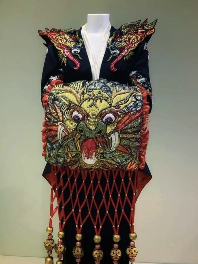 a fulllength shot of a traditional asian costume, possibly from a play or ceremony, is displayed on a mannequin. the garment features intricate embroidery, particularly a large, stylized dragon head on the front. the shoulders are adorned with similar dragon motifs. the lower portion includes a fringe of red cords and decorative gold beads. the costume is primarily dark in color, contrasting with the vibrant embroidery and gold embellishments. the background is a plain, lightcolored wall.