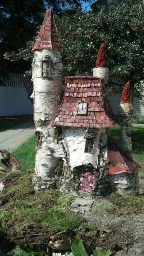 a whimsical miniature fairy house is nestled in a garden setting. the house is constructed primarily from birch bark, giving it a rustic and natural appearance. it features a tall tower with a coppercolored pointed roof. the main house has a gabled roof covered in individual shingles, also in shades of red and brown. there are small windows and a tiny door, adding to the enchanting feel. the house is surrounded by moss, plants, and other garden elements, creating a magical scene.