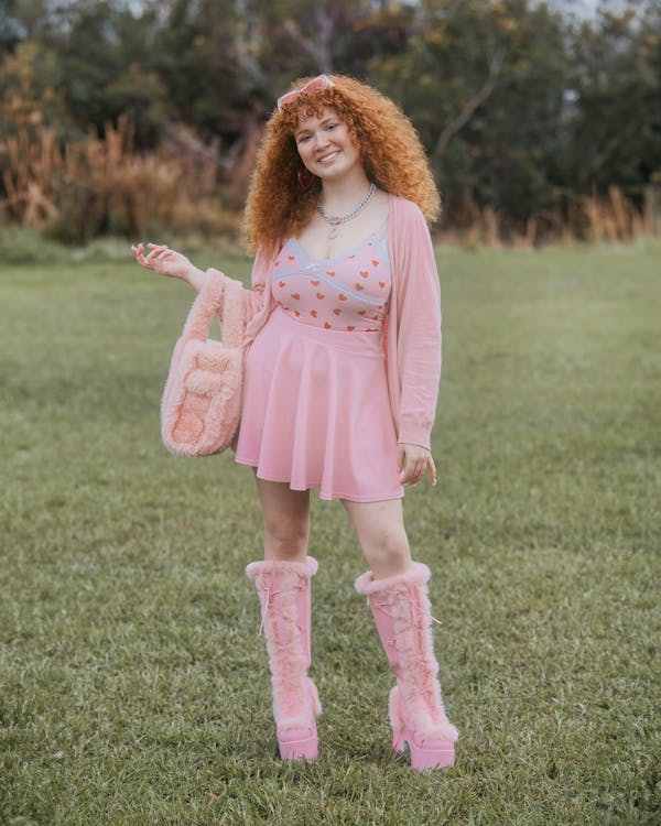 a young redhead woman with curly hair is standing in a grassy park. she's wearing a cute pastel pink outfit: a pink heartprinted cami, a pink skirt, and a pink cardigan. she's also wearing fluffy pink platform boots and carrying a fluffy pink purse. she's smiling happily at the camera. the overall style is cute and kawaii.